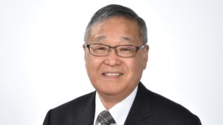 Smiling middle-aged man wearing glasses, a dark suit, and a plaid tie, photographed against a white background.