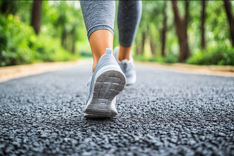 Close-up of a person wearing gray running shoes walking on a paved park path with greenery around.