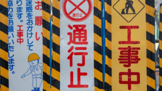 Barrier with multiple construction signs: central red '通行止' (no through traffic), flanked by yellow '工事中' (work in progress) panel and a left panel showing a construction worker and warning text in Japanese.