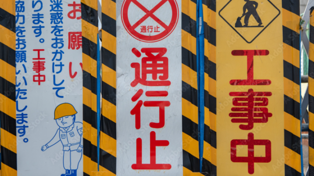 Barrier with multiple construction signs: central red '通行止' (no through traffic), flanked by yellow '工事中' (work in progress) panel and a left panel showing a construction worker and warning text in Japanese.