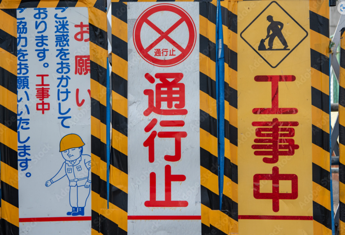 Barrier with multiple construction signs: central red '通行止' (no through traffic), flanked by yellow '工事中' (work in progress) panel and a left panel showing a construction worker and warning text in Japanese.