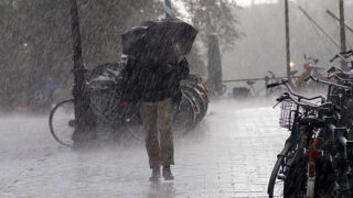 Person walking with a large umbrella through heavy rain on a wet city street, bicycles parked along the curb.