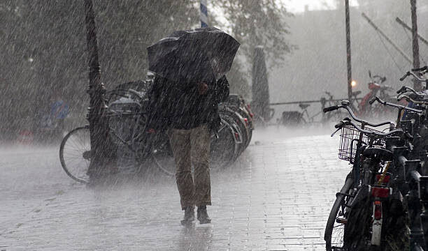Person walking with a large umbrella through heavy rain on a wet city street, bicycles parked along the curb.